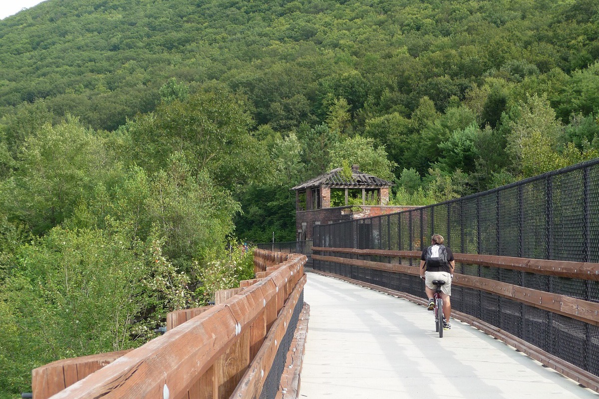 A timber and steel rail trail bridge running through the Lehigh gorge, dense green woodland climbing the hillside beyond, a ruined brick building at the far end and a cyclist riding away down the deck.