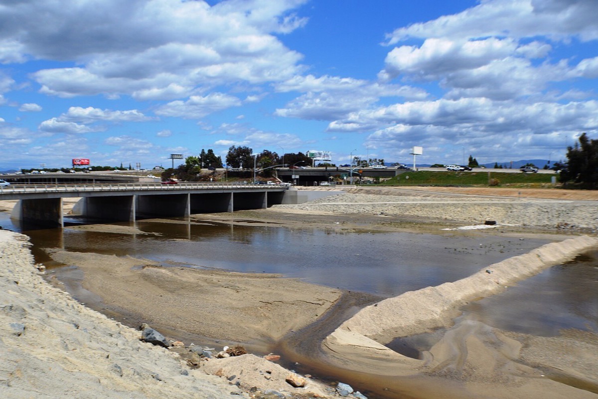 The concrete river channel running through Anaheim under a road bridge, a shallow braided stream crossing pale sand at the bottom of it and tall white cloud above.