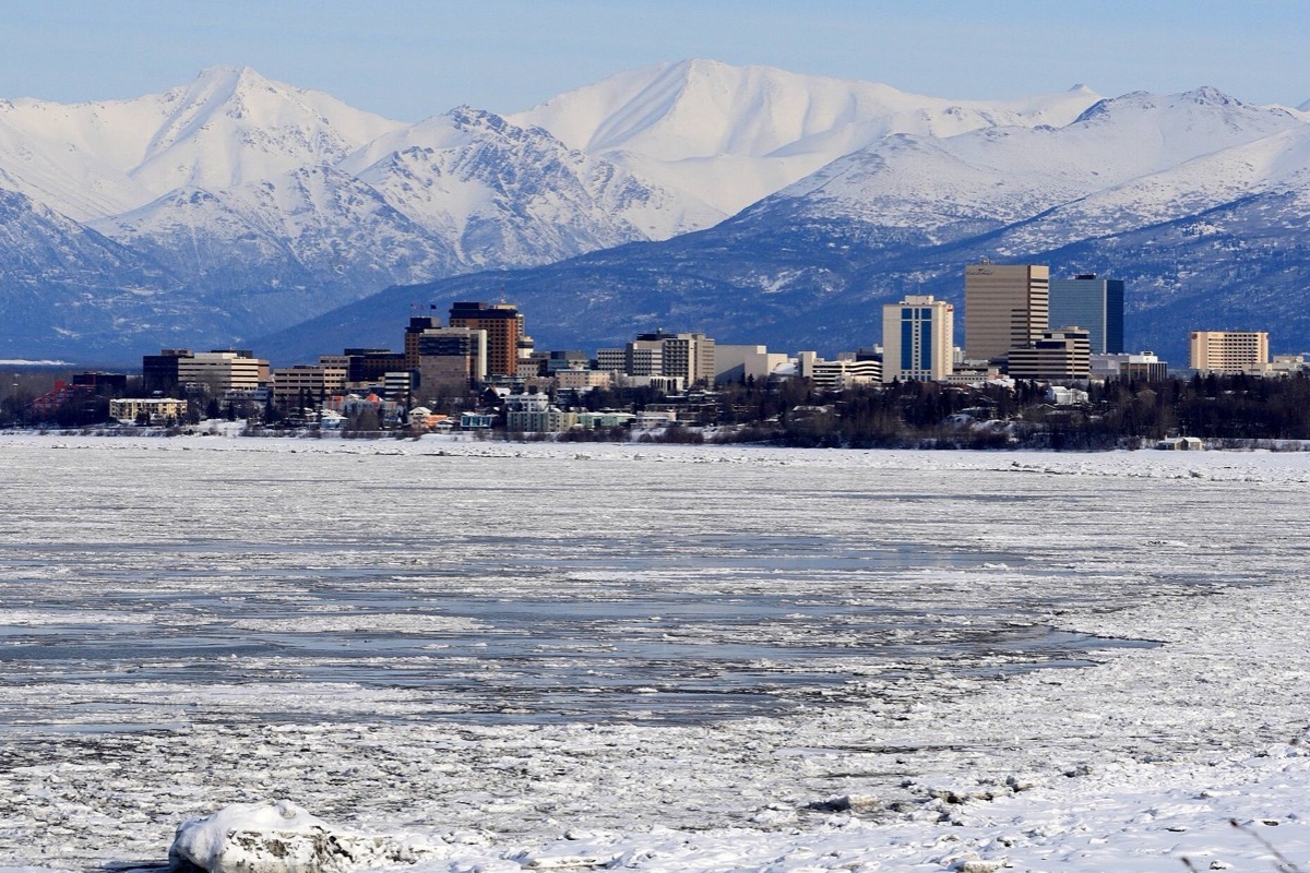 The Anchorage skyline low along the far shore of a frozen inlet, broken ice filling the whole foreground and the snow covered Chugach range standing behind the city.