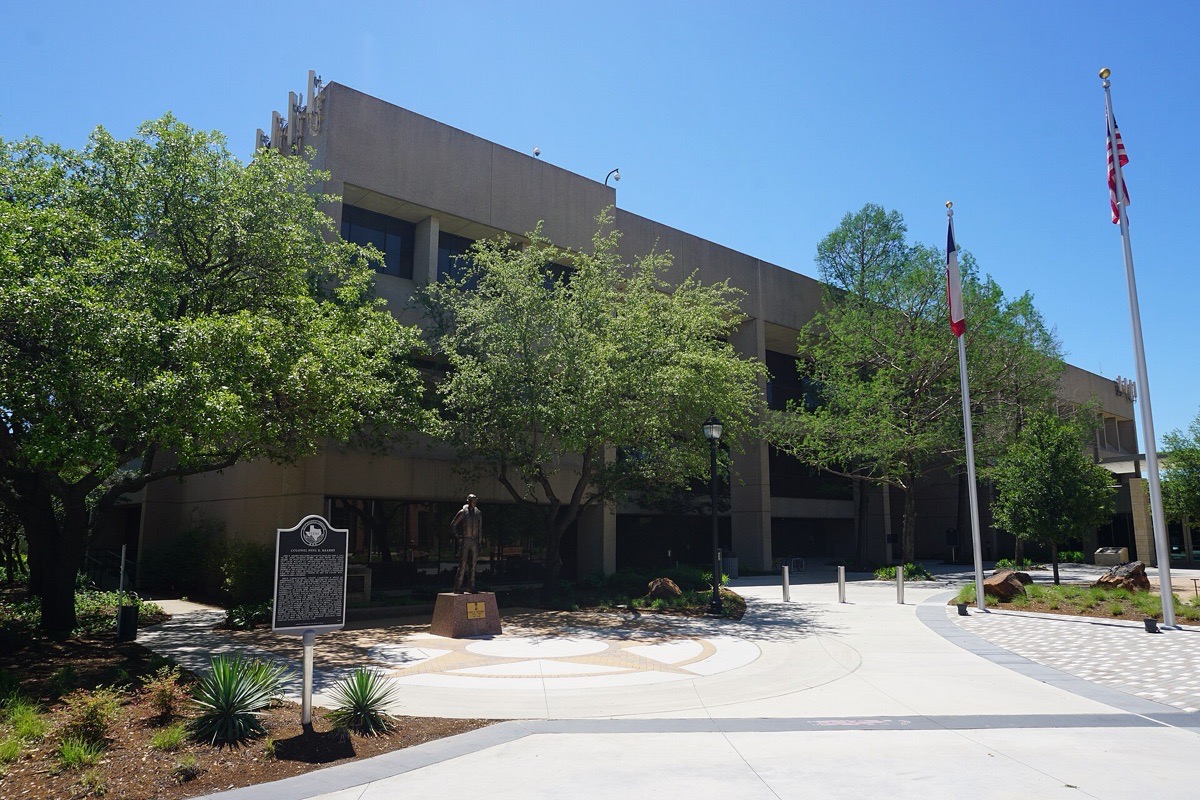 Arlington civic buildings in Texas, a low concrete and glass block behind mature green trees with a paved forecourt, a historical marker and two flagpoles in front under a clear blue sky.