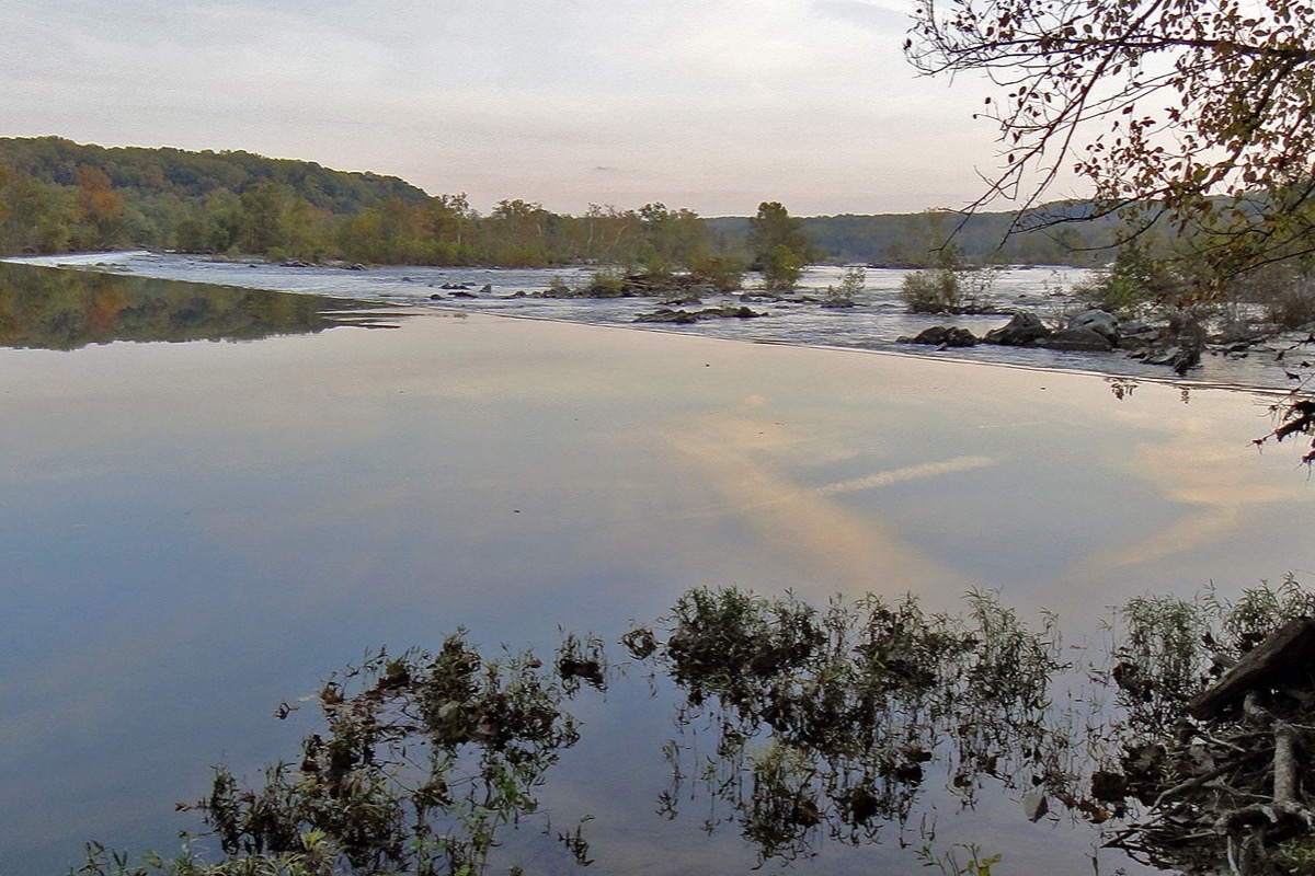 The Potomac upstream of Washington, still water reflecting cloud, a long low dam line running across the river and autumn woods closing the far bank.