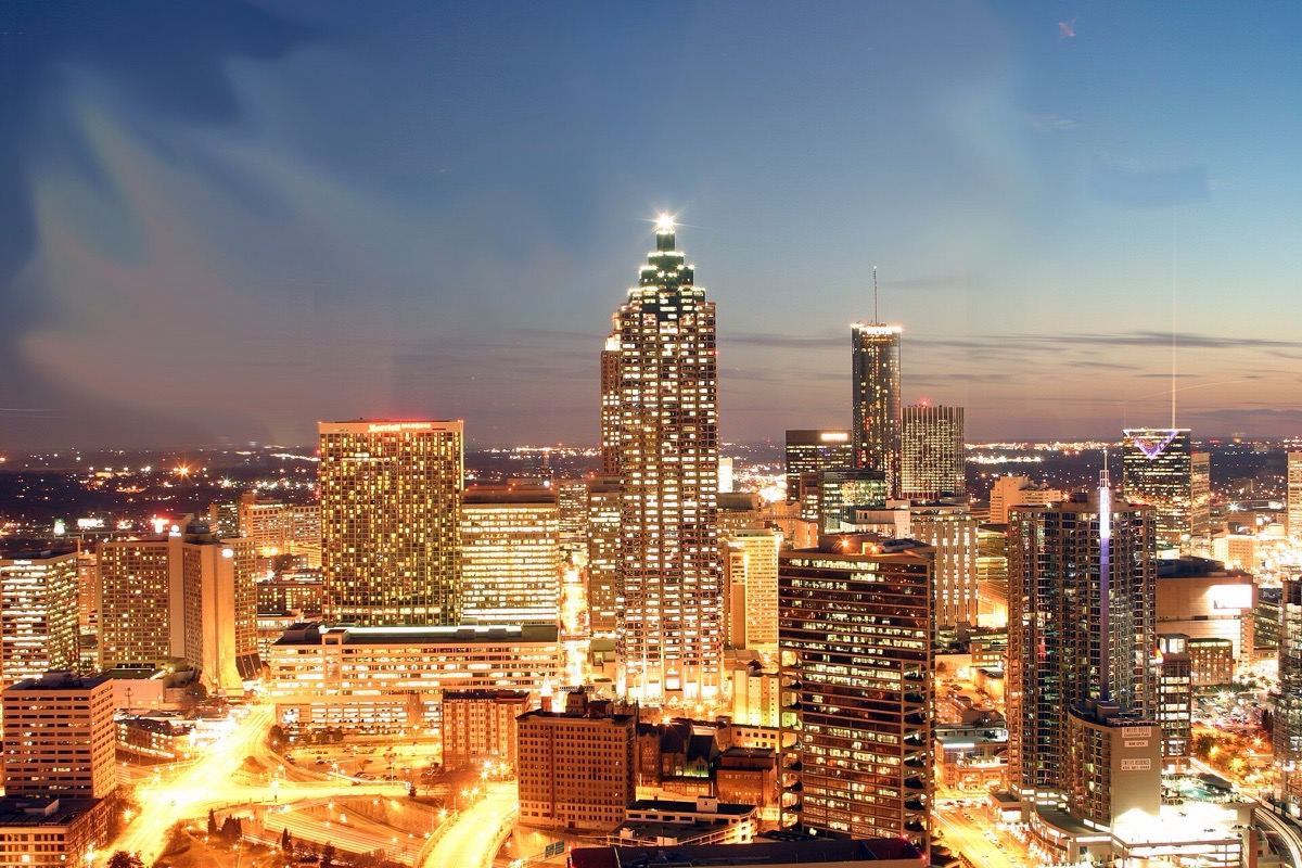 The Atlanta skyline at dusk, office towers lit from within and a lattice of street lights running away below them under a deep blue sky.