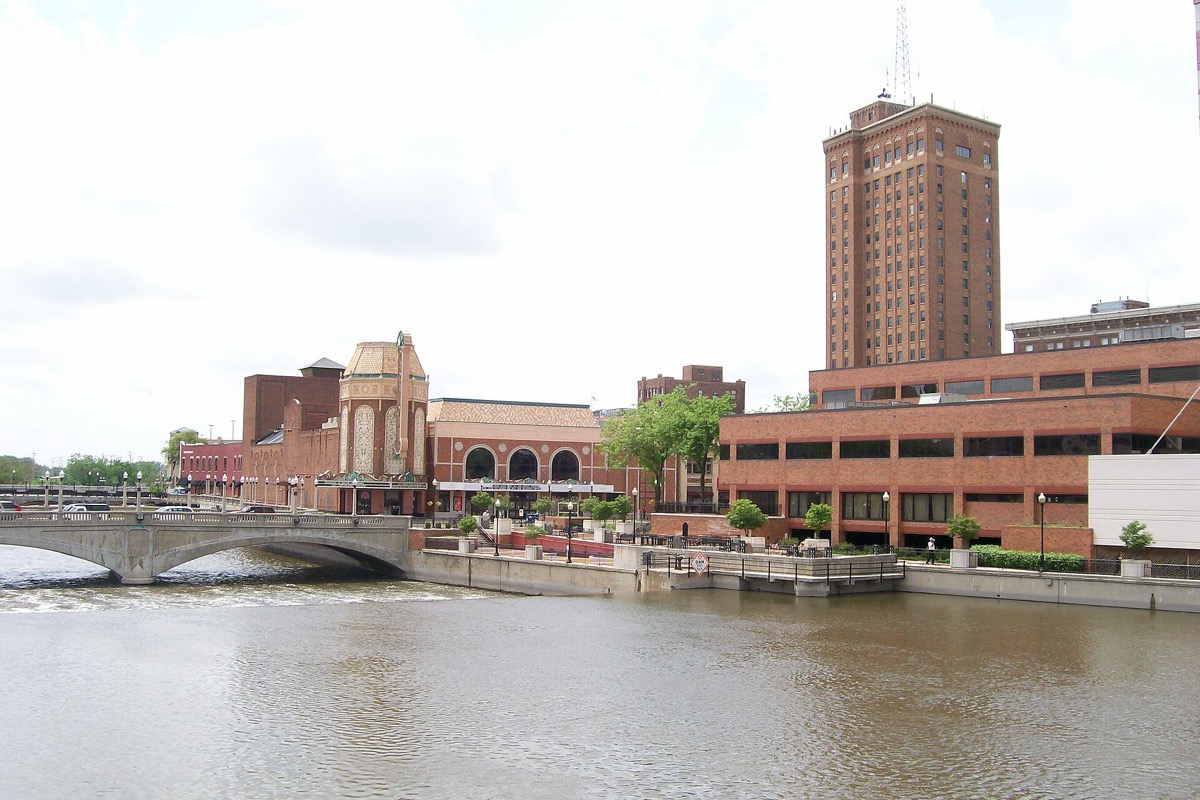 The Fox River through the middle of Aurora, a stone arch bridge crossing on the left, an ornate theatre front and a tall brick tower along the far bank, and a concrete river walk at the water's edge.