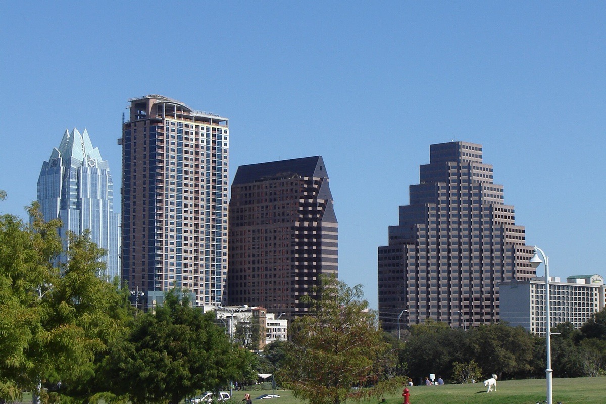 Austin office and apartment towers standing above a broad green lawn and a line of mature trees, one of them in blue glass, under a clear Texas sky.
