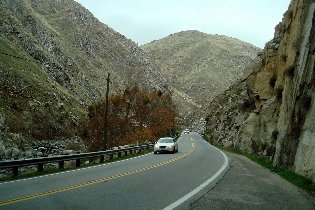 A two lane road curving through the Kern canyon above Bakersfield between a guardrail and a face of tilted rock, with a car coming down and dry hills rising on both sides.