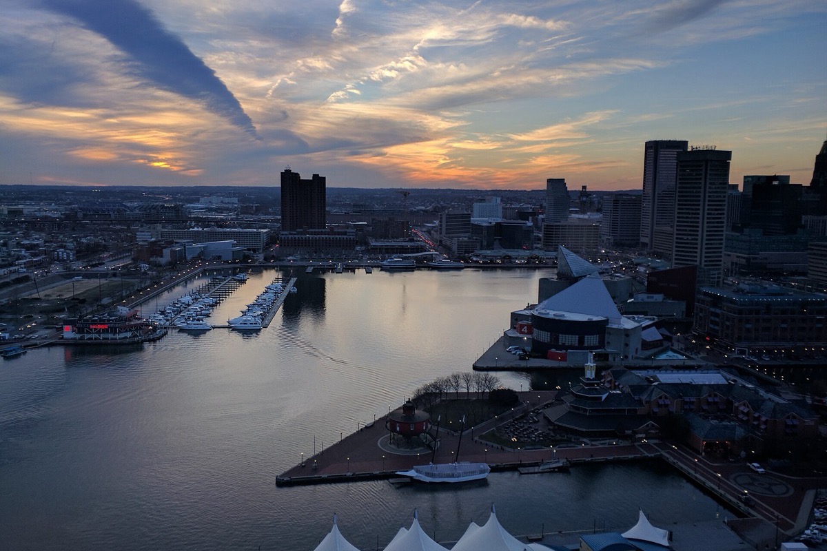 Baltimore harbour from above at sunset, the water still between the piers, moored boats along the near side, the office towers catching the last light and a bank of cloud lit orange behind them.