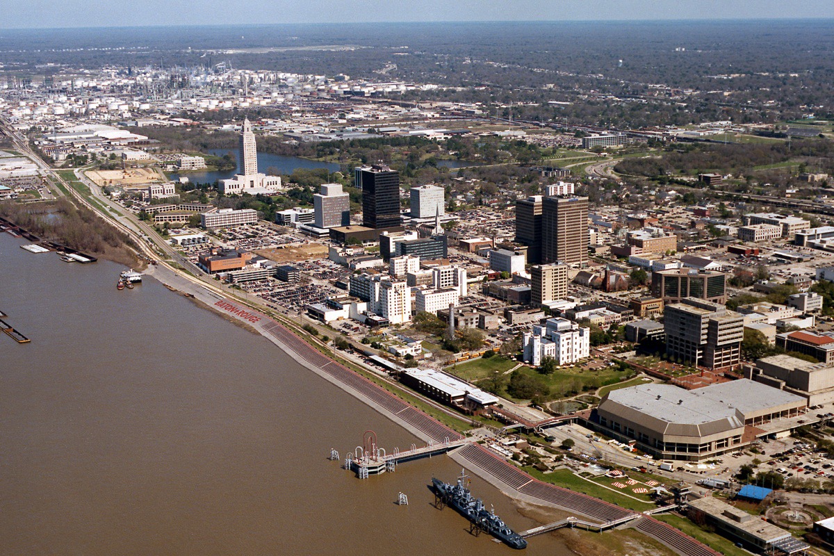 An aerial view of Baton Rouge along the Mississippi, the levee and river walk in the foreground, the downtown and the capitol tower behind it, and industrial plant stretching along the bank upriver.