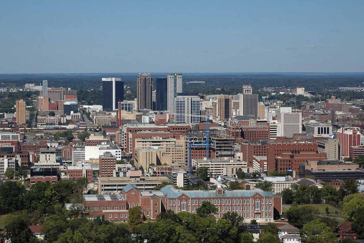 Birmingham, Alabama seen from a wooded ridge above the valley: a compact cluster of mid-rise office towers on the flat ground, low red brick blocks and a construction crane in front of them, and tree-covered hills closing the horizon under a clear blue sky.