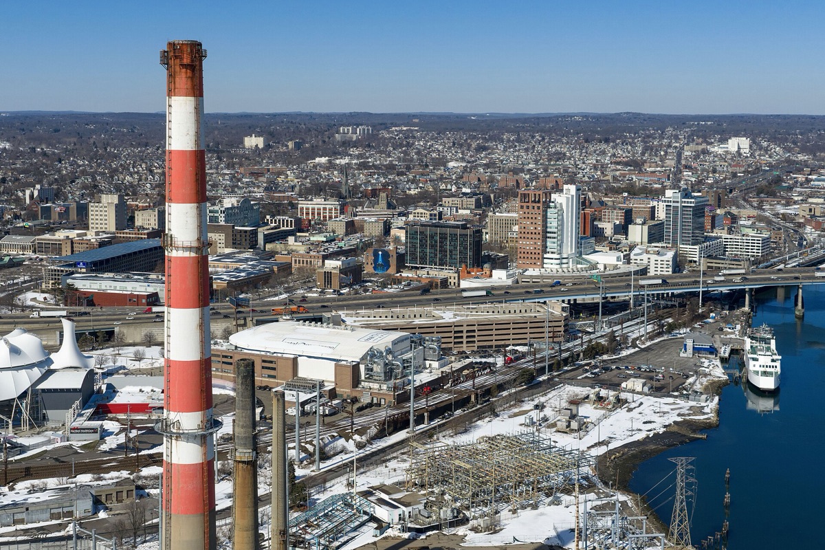 Bridgeport from the air under snow, a tall red and white banded power station chimney rising in the foreground, rail yards, an electrical substation and a ferry berth along the harbour, with the city and low wooded hills behind.