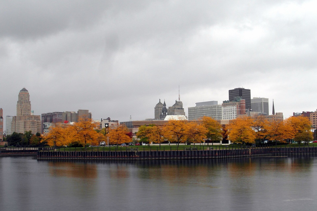 The Buffalo skyline seen across the still water of the Erie Basin marina in October, a line of trees in full autumn colour along the near shore and the office towers standing behind them.