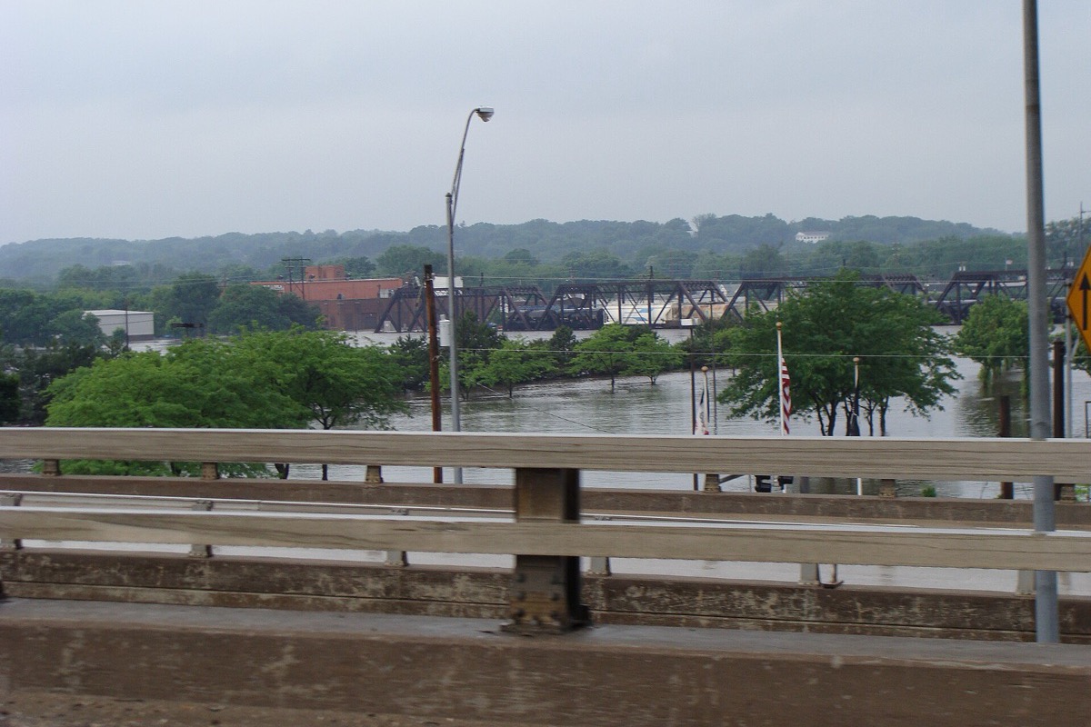 The Cedar River out of its banks in June 2008, brown water standing among riverside trees and reaching the deck of a steel railway truss bridge, photographed from a road bridge with its concrete guardrail across the foreground.