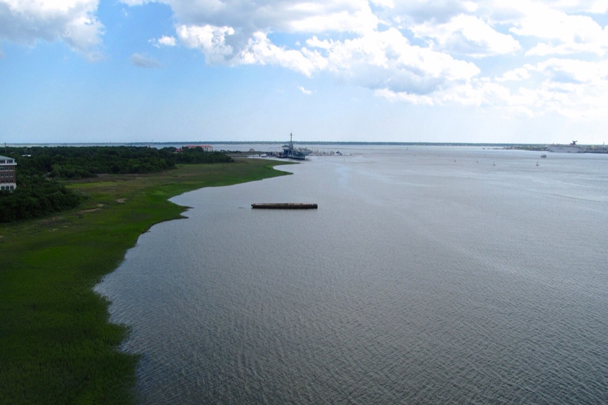 Charleston harbour from above, a wide sheet of grey green water with salt marsh and a wooded bank on the left, a moored barge mid channel and a ship at a pier in the distance, under tall summer cloud.