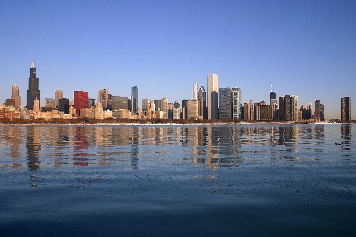 The Chicago skyline seen low across Lake Michigan on a calm morning, the whole line of towers standing above a pale shoreline and mirrored in flat water.