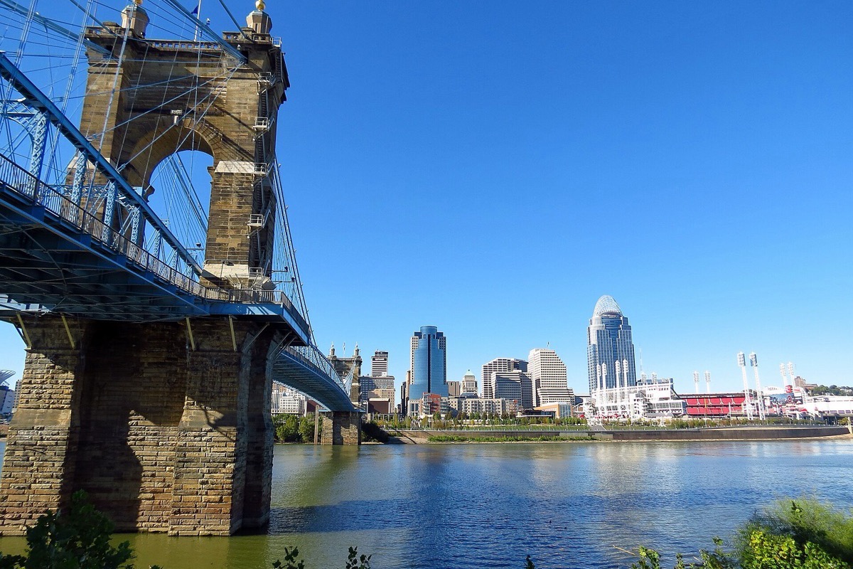 One stone tower and the cables of the Cincinnati suspension bridge in the foreground, the river below and the city skyline standing along the far bank under a deep blue sky.