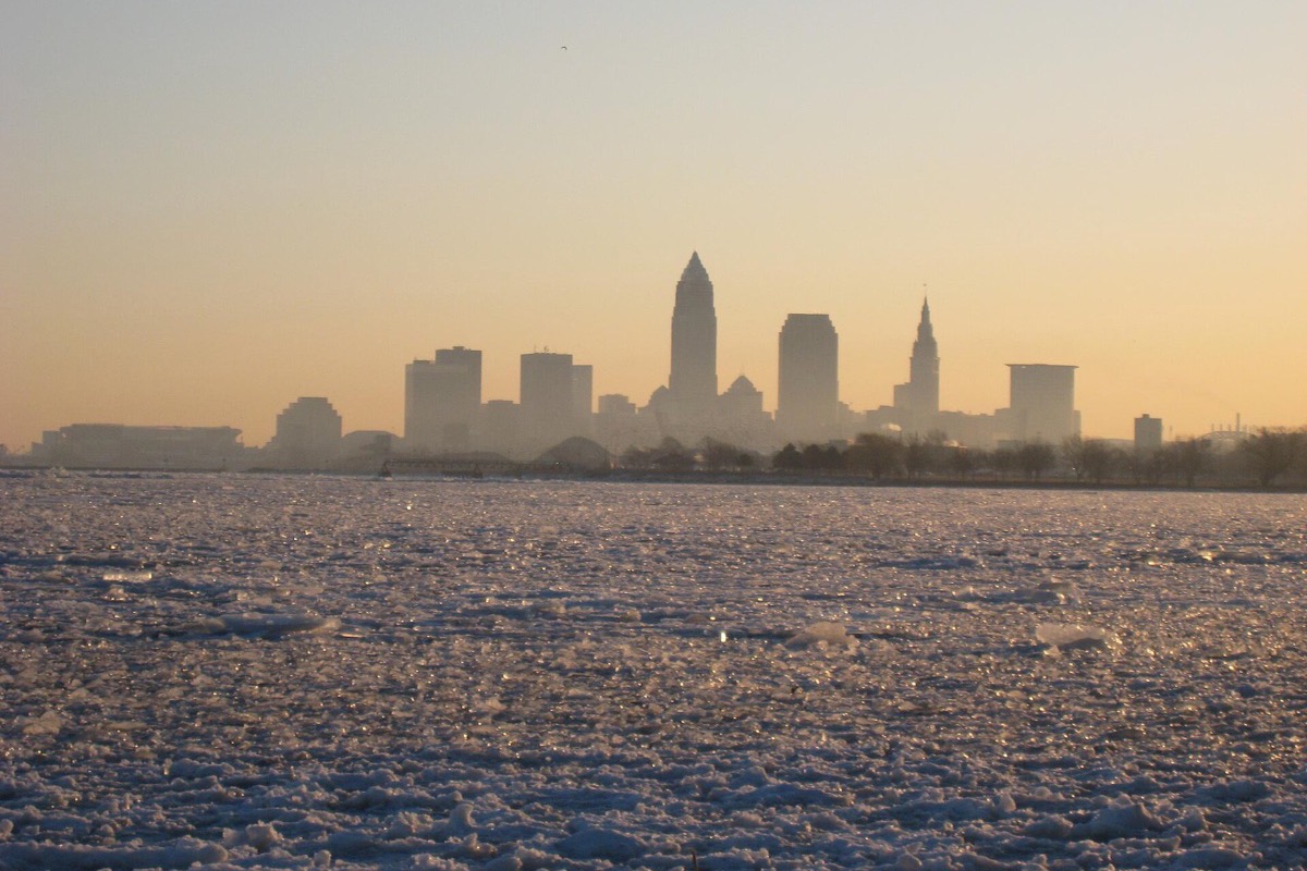 The Cleveland skyline in silhouette across a completely frozen Lake Erie, broken pale ice filling the whole foreground and low winter sun turning the haze gold.