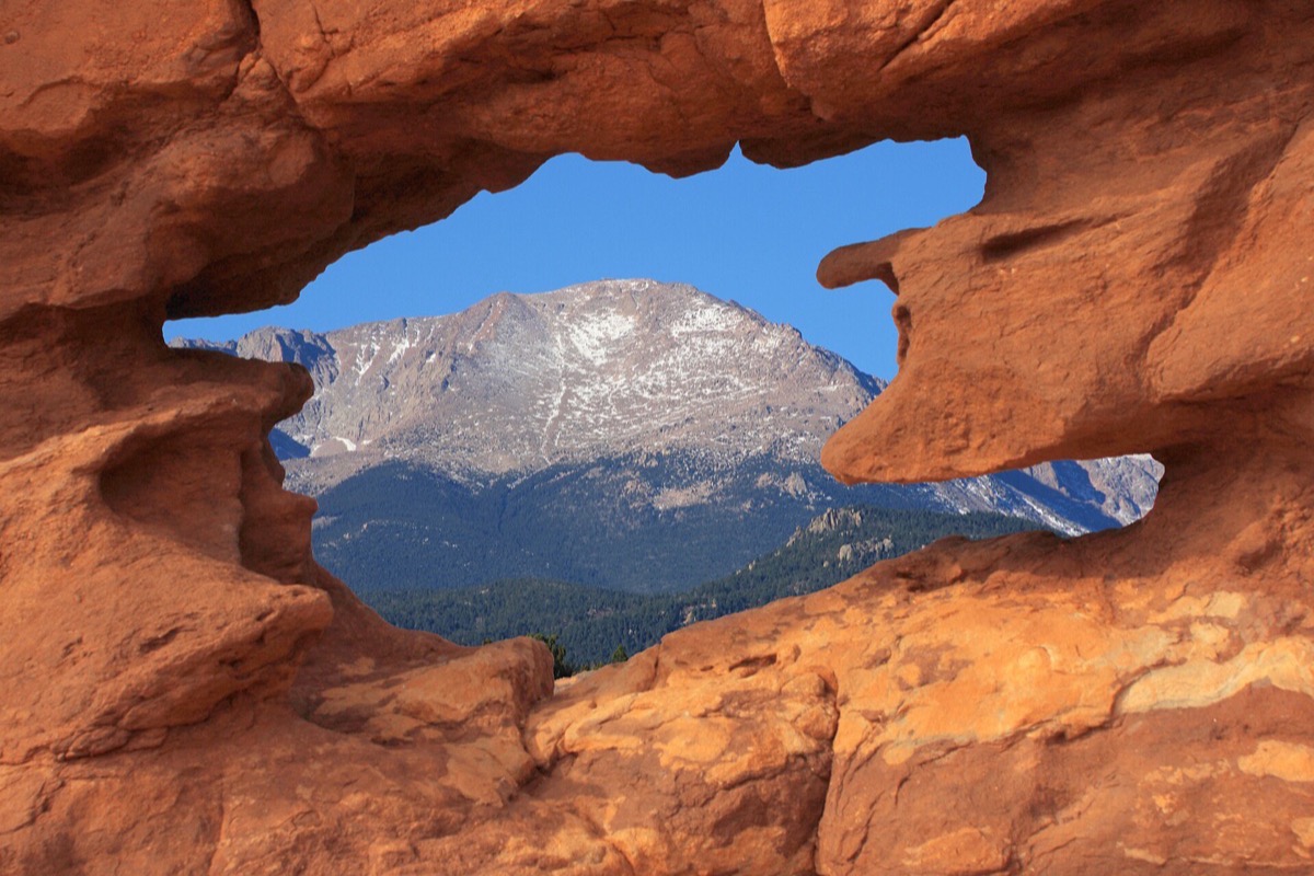 The snow covered summit of Pikes Peak framed through a natural window in a red sandstone fin, the rough rock filling the frame around the opening.