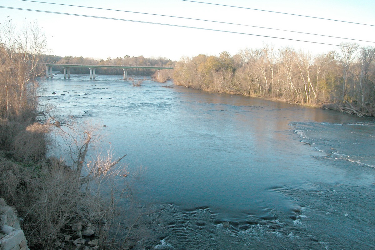 A broad shallow river running over rock shoals under a low concrete road bridge, bare winter trees along both banks and power lines crossing the top of the frame.