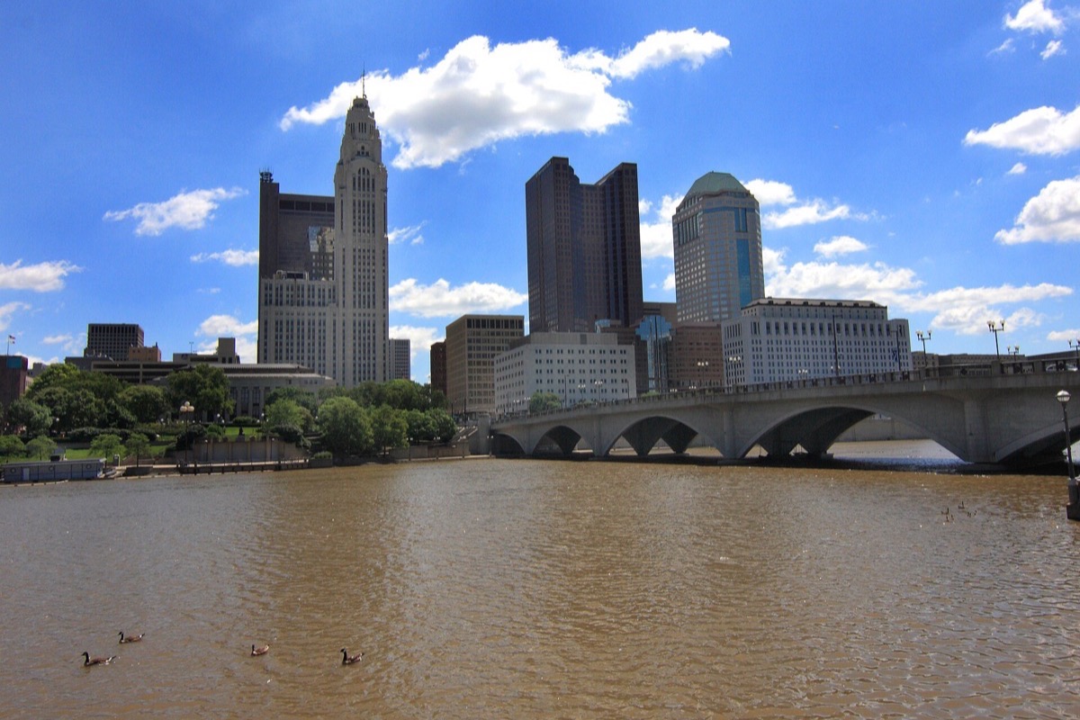 The Columbus skyline standing above the Scioto, a low arched bridge crossing the brown water in front of it and tall white cloud running across a blue sky behind.