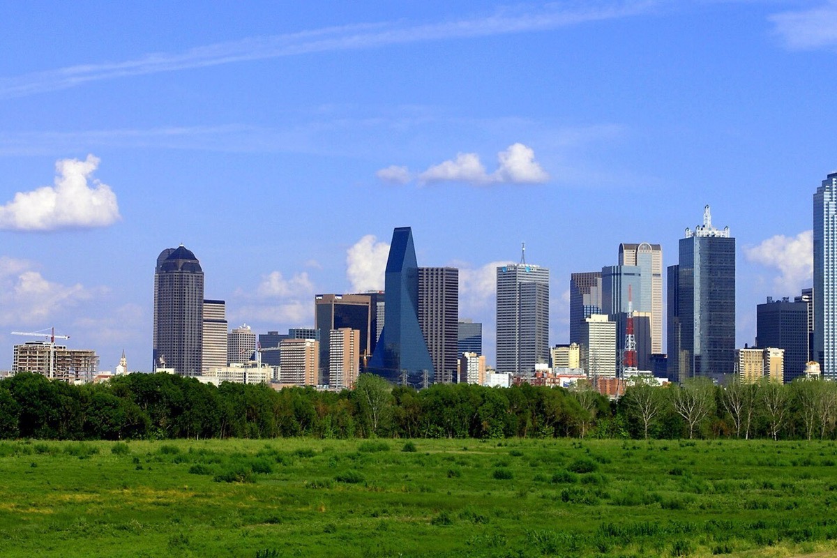 The Dallas skyline seen across the green floodplain below it, a line of trees between, and a tall bank of white cloud standing behind the towers.