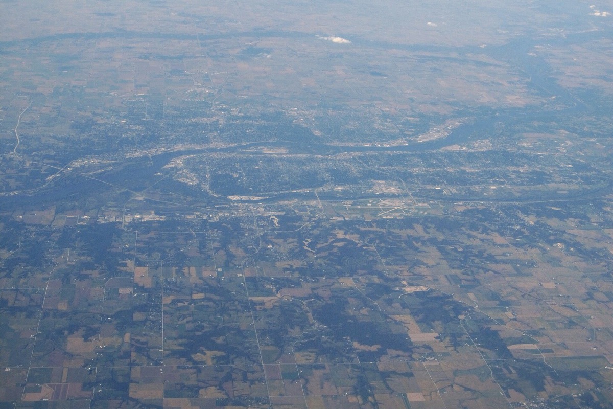 A high aerial view of the Quad Cities, the Mississippi curving through the middle of the frame with built-up streets on both banks and a grid of farmland running out to a hazy horizon.