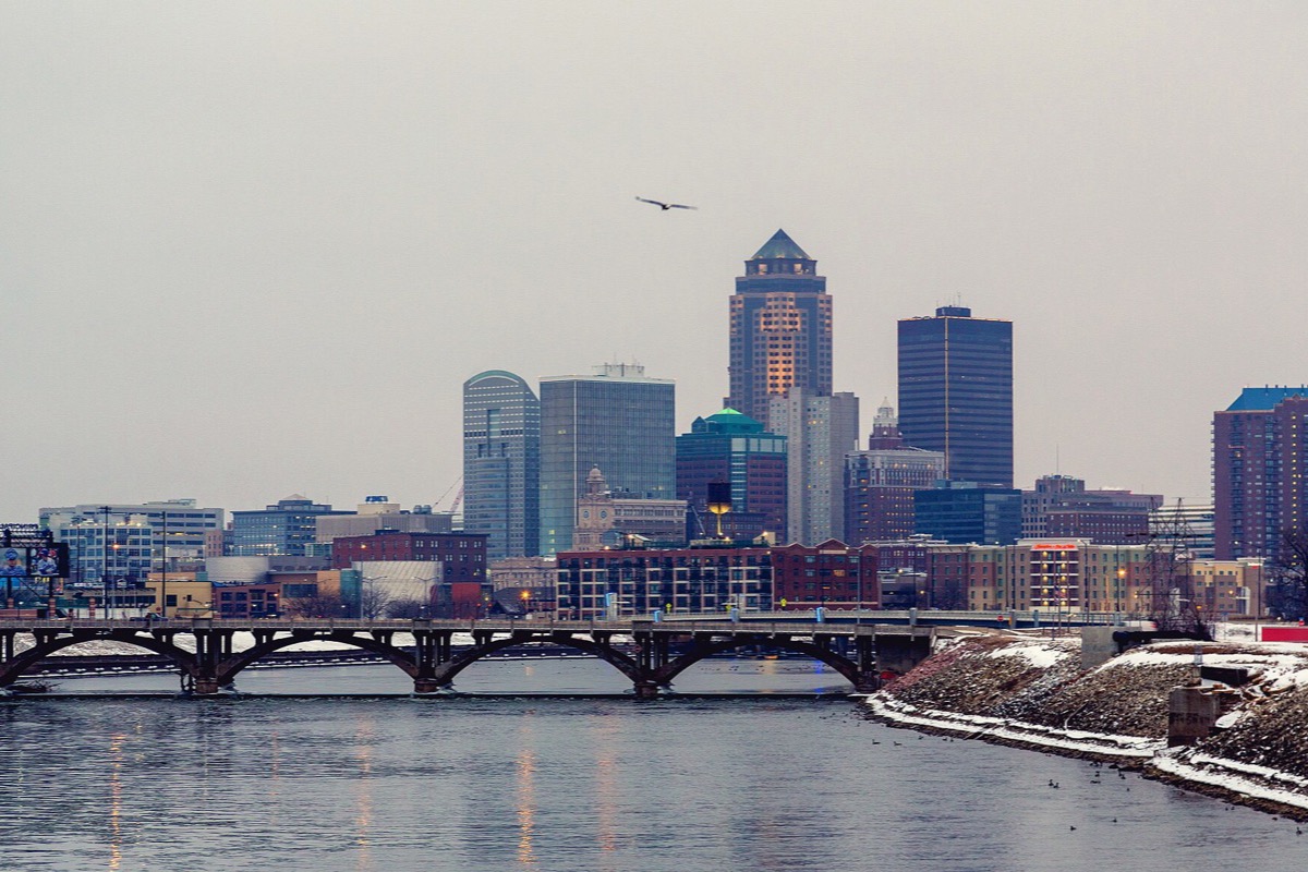 Downtown Des Moines across the river on a grey winter afternoon, a low arched road bridge spanning dark water in the foreground, snow lying along the far bank, and a cluster of office towers rising behind.
