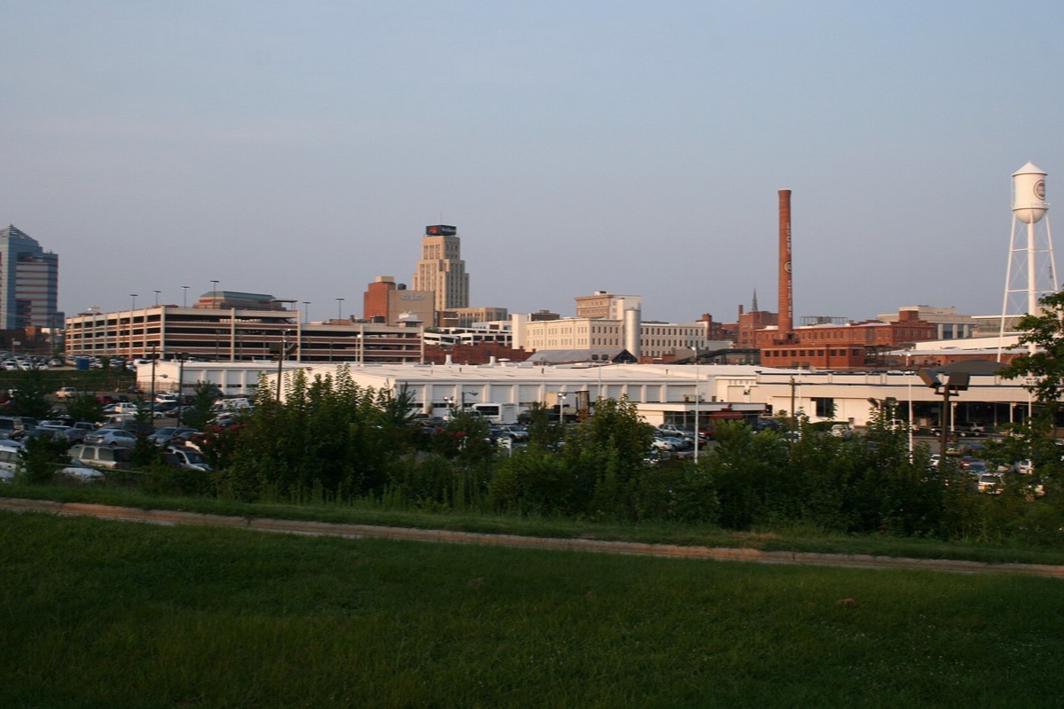 The Durham skyline on a bright morning, a tall brick smokestack and a white water tower standing among the office blocks, with parked cars and green grass in front.
