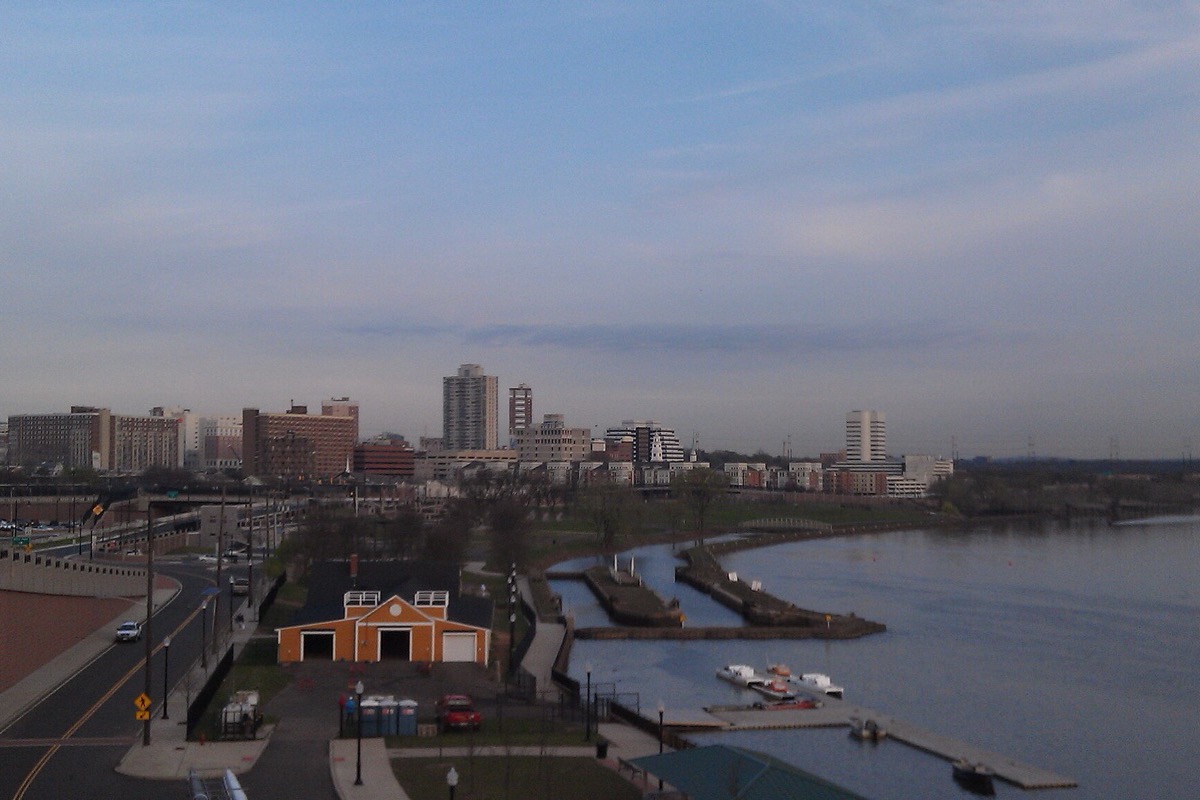 The Raritan River at dusk, a low orange boathouse and a floating dock on the near bank, a landscaped riverfront park beside a road, and a mid-rise skyline along the far shore.