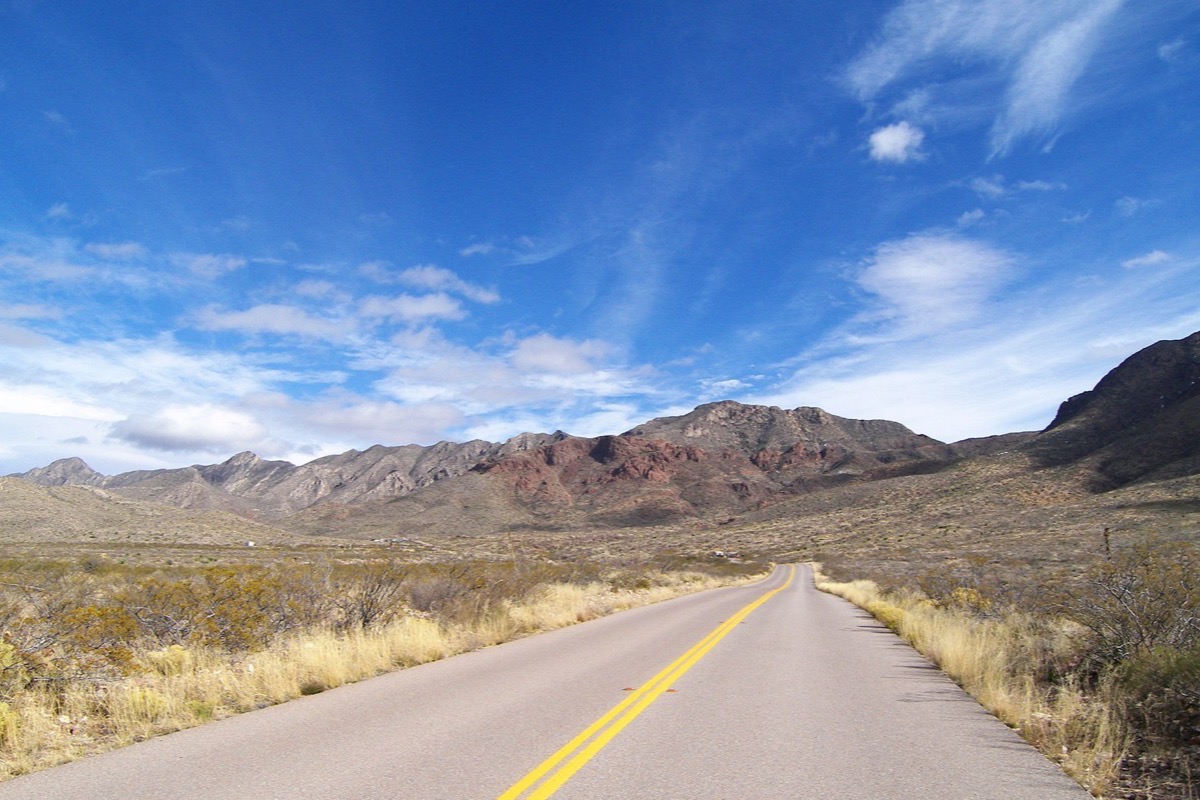 A two lane road running straight toward the Franklin mountains above El Paso, dry yellow grass on both shoulders and the bare red and grey range standing across the horizon under a blue sky.