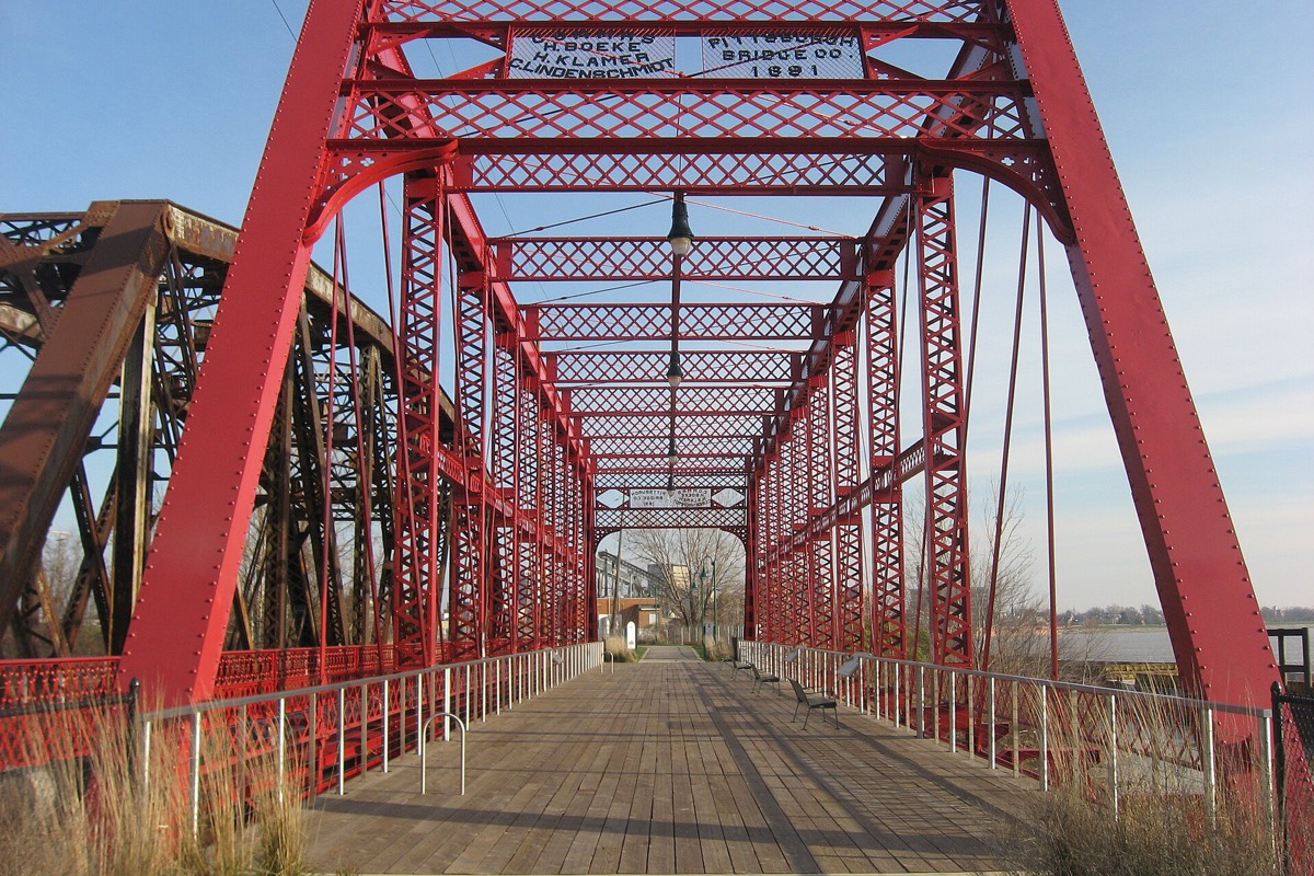 Looking along the timber deck of a restored red iron truss bridge, its lattice framework arching overhead, a builder's plate fixed to the portal and the river visible to the right.