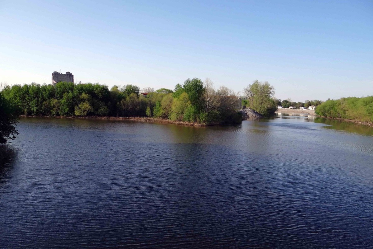 Two rivers meeting at Fort Wayne on a clear spring morning, a wooded point dividing the channels, green banks on both sides and a single tall building above the treeline.