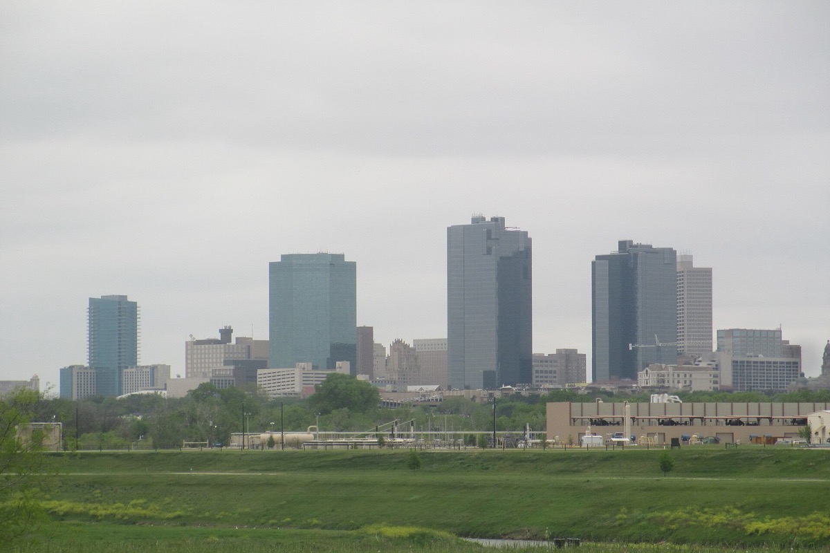 The Fort Worth skyline in soft haze beyond a wide strip of green floodplain, the towers pale grey and the whole view flat and open.