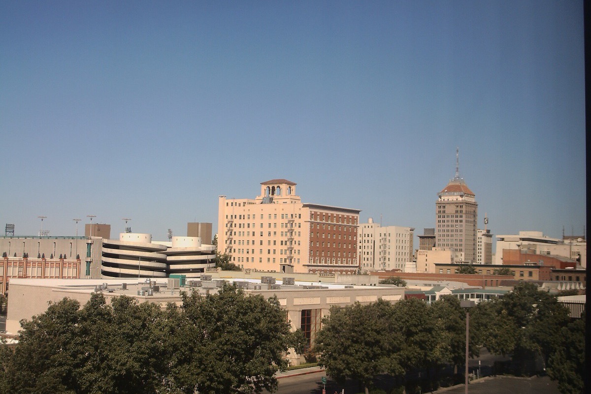 The Fresno skyline seen over a line of mature trees, an ornate tiled tower standing among the older stone blocks and newer offices under a deep blue sky.