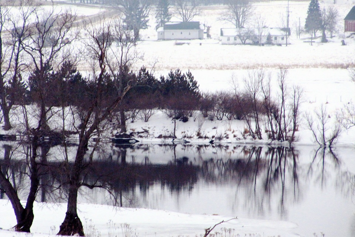 The Grand River in winter west of Grand Rapids, snow lying along both banks, bare trees standing in still grey water and farm buildings on the rise beyond.