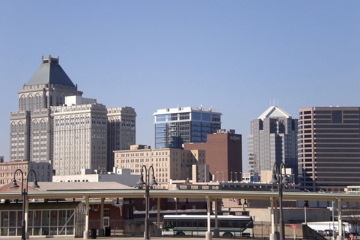 The Greensboro skyline behind a rail cut, older stone faced buildings and newer glass ones standing together, with a bus stopped on the near side.