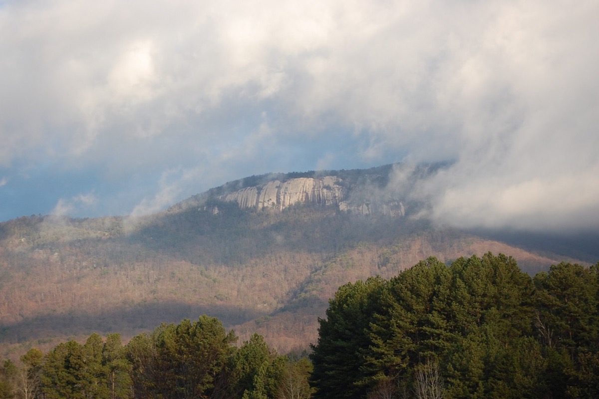 A granite dome on a forested ridge above the Upstate, its bare rock face catching light through low cloud, with winter woodland running down the slope and pines in the foreground.