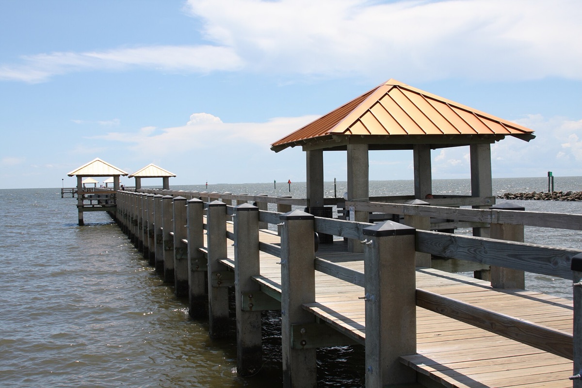 A timber fishing pier running out over calm Gulf water, with copper roofed shelters along it and a rock breakwater on the horizon, under a blue sky with light cloud.