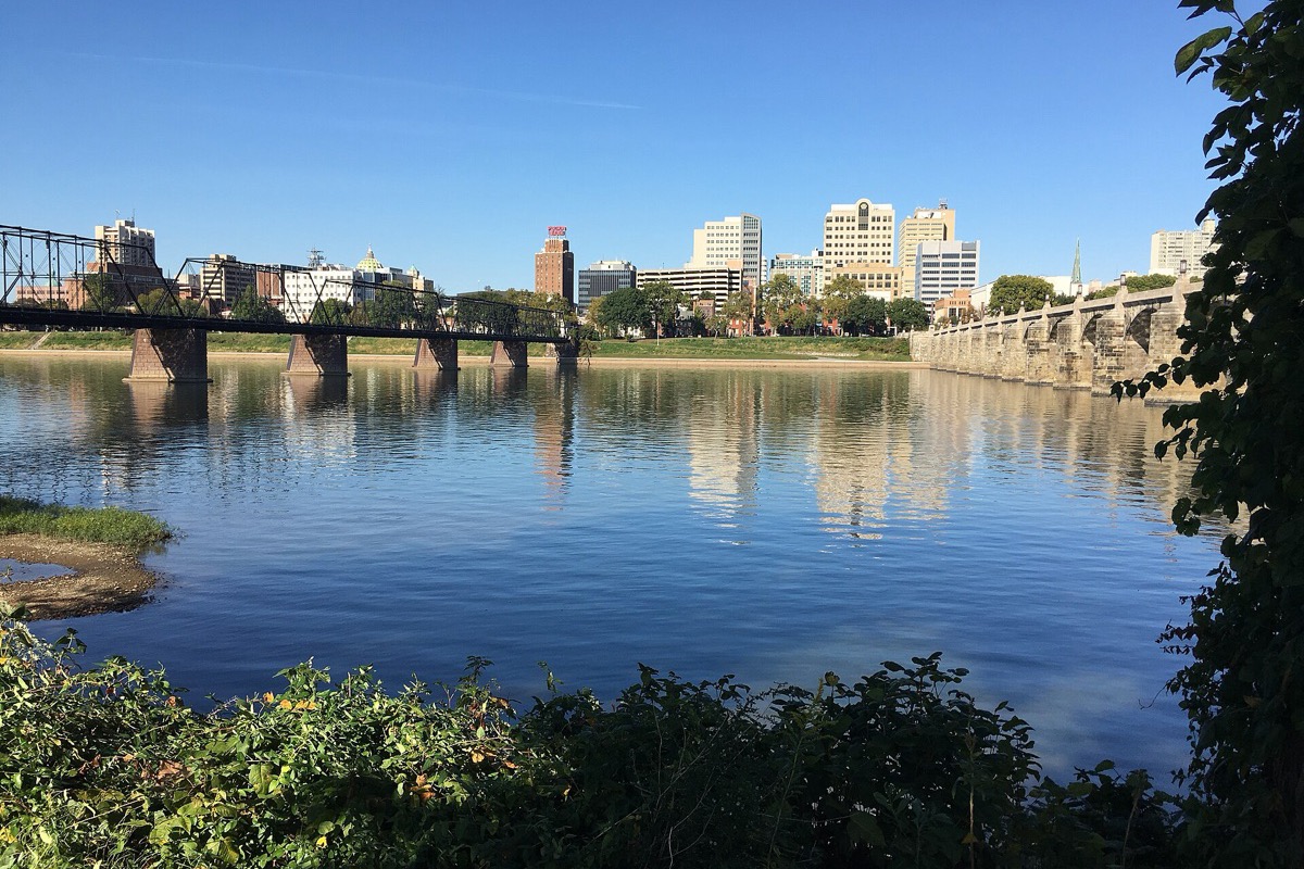 The Susquehanna at Harrisburg on a clear autumn day, a black steel truss bridge and a long stone arch bridge crossing the river, the capitol dome and the city skyline reflected in still water.