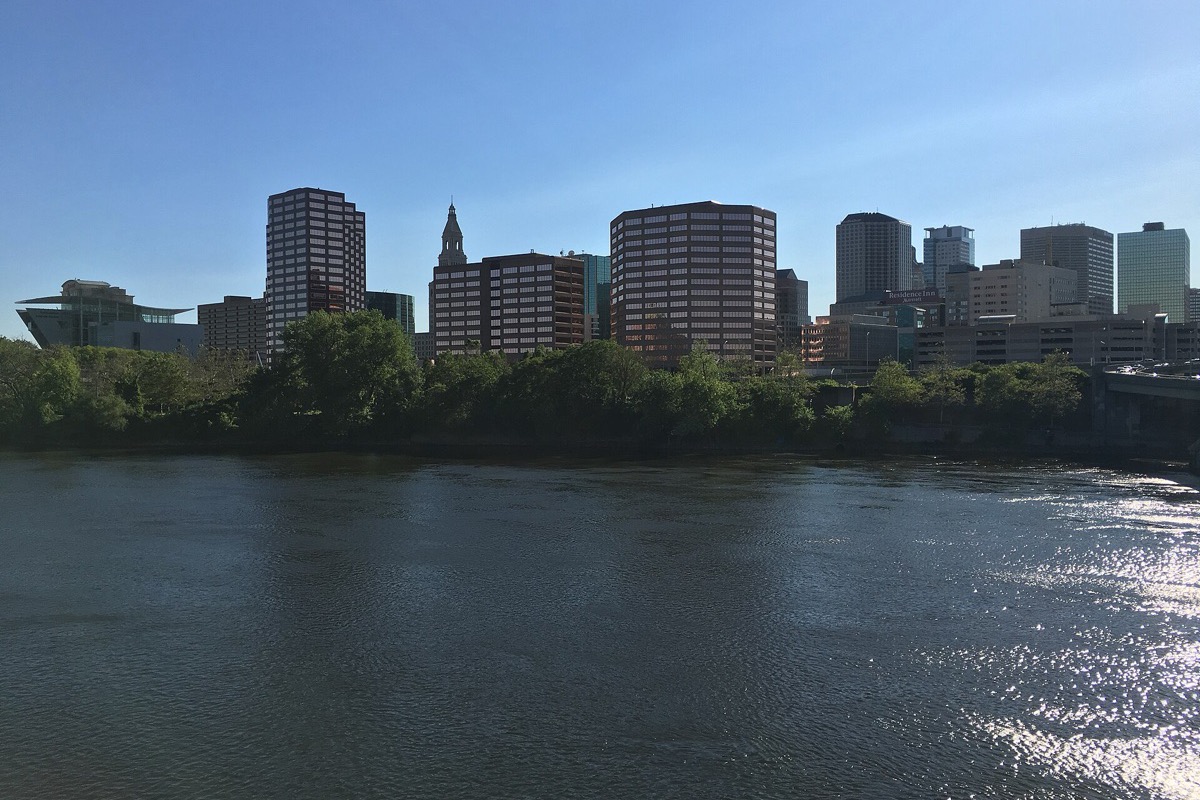 The Connecticut River in front of the Hartford skyline on a clear summer afternoon, a wooded bank between the water and the office towers, and sunlight scattering off the surface across the foreground.