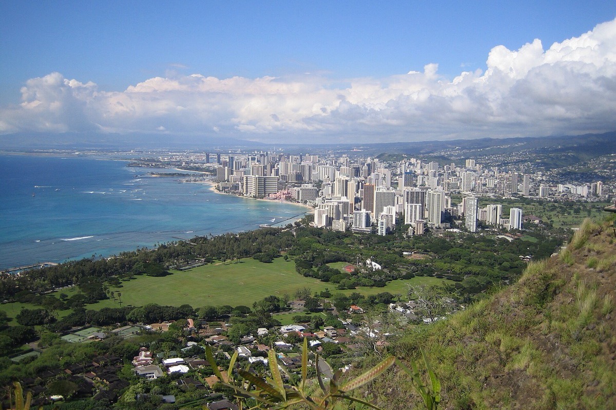 Honolulu and the Waikiki shoreline seen from the crater rim above it, a long curve of hotels along the beach, green parkland in the middle and the ocean running out to a cloud bank.