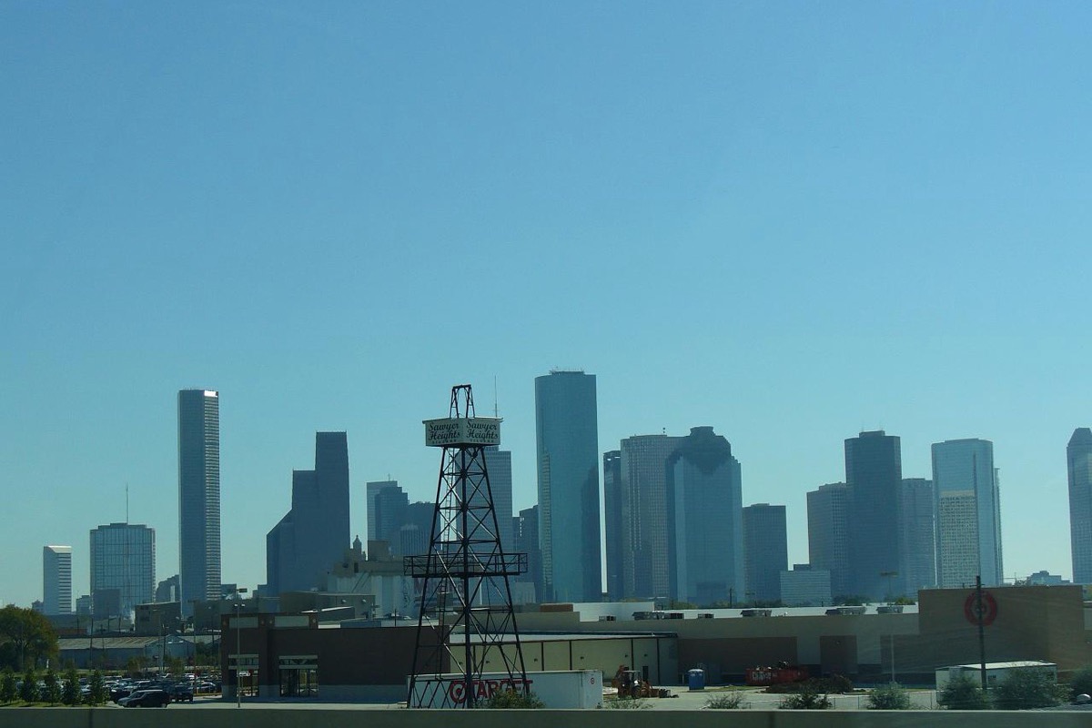 The Houston skyline under a clear sky with an old steel oil derrick standing in the foreground, low industrial buildings between it and the towers.