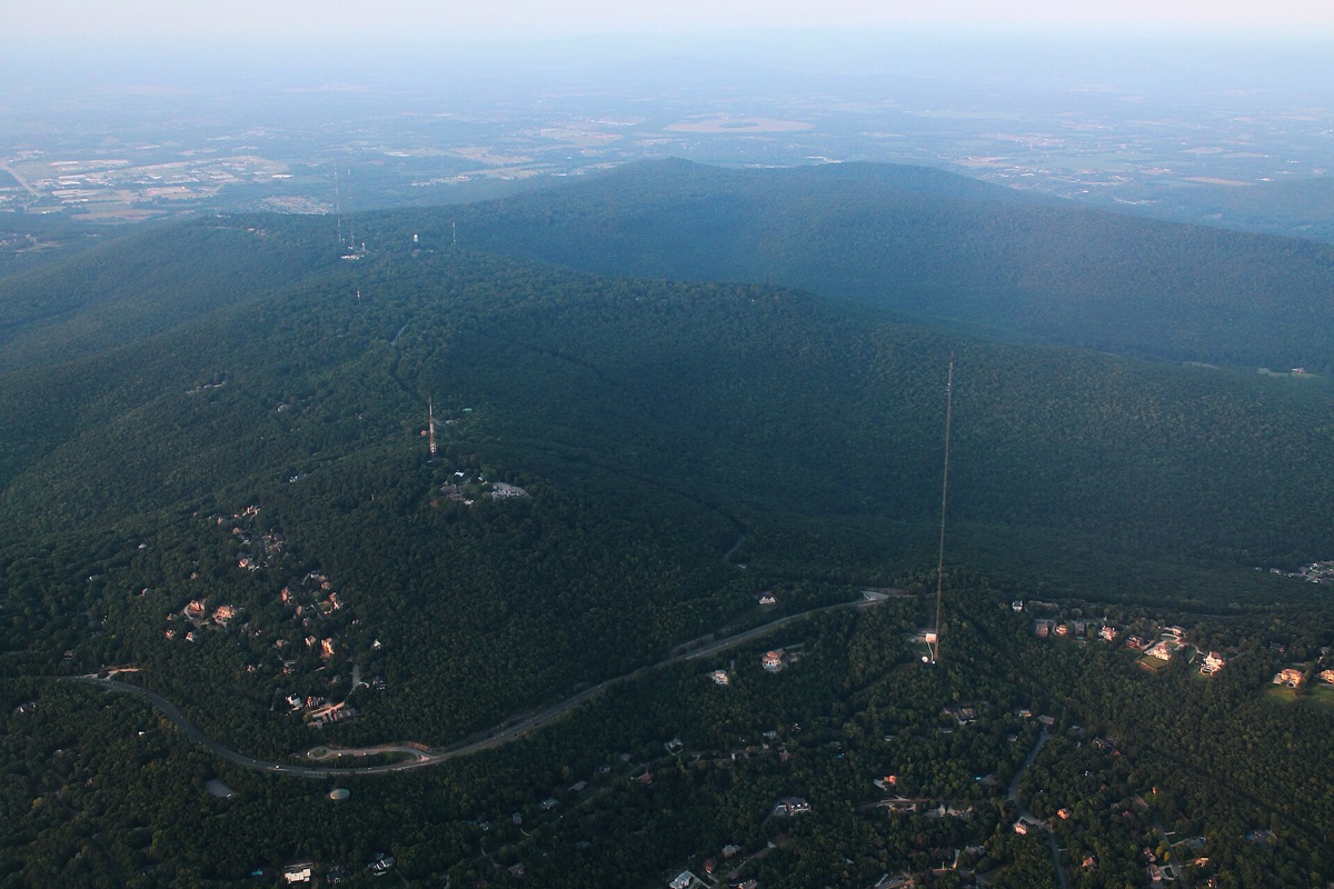 Aerial view of the wooded ridge above Huntsville, Alabama, with tall slender transmission masts rising out of dense tree cover, houses tucked along a winding ridge road, and the hazy Tennessee Valley stretching flat to the horizon.