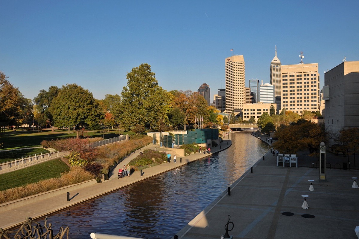 The canal in Indianapolis in autumn, still water between stone walls with a paved walkway along one side, yellow and orange trees on the bank and the city towers rising behind them.