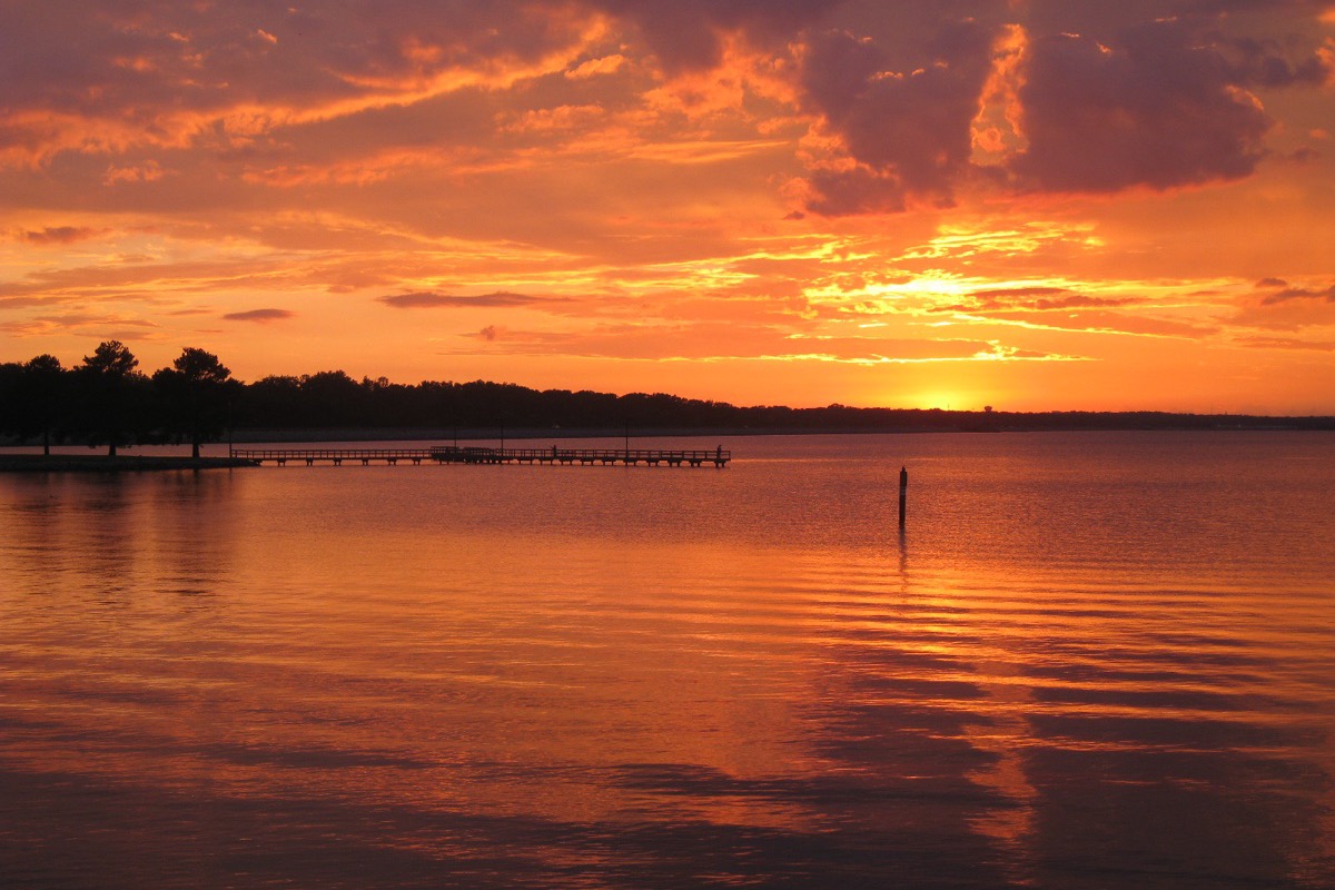An orange sunset over a wide reservoir, banked cloud lit from below, a low jetty and a dark treeline silhouetted on the far shore, and the colour reflected across still water in the foreground.