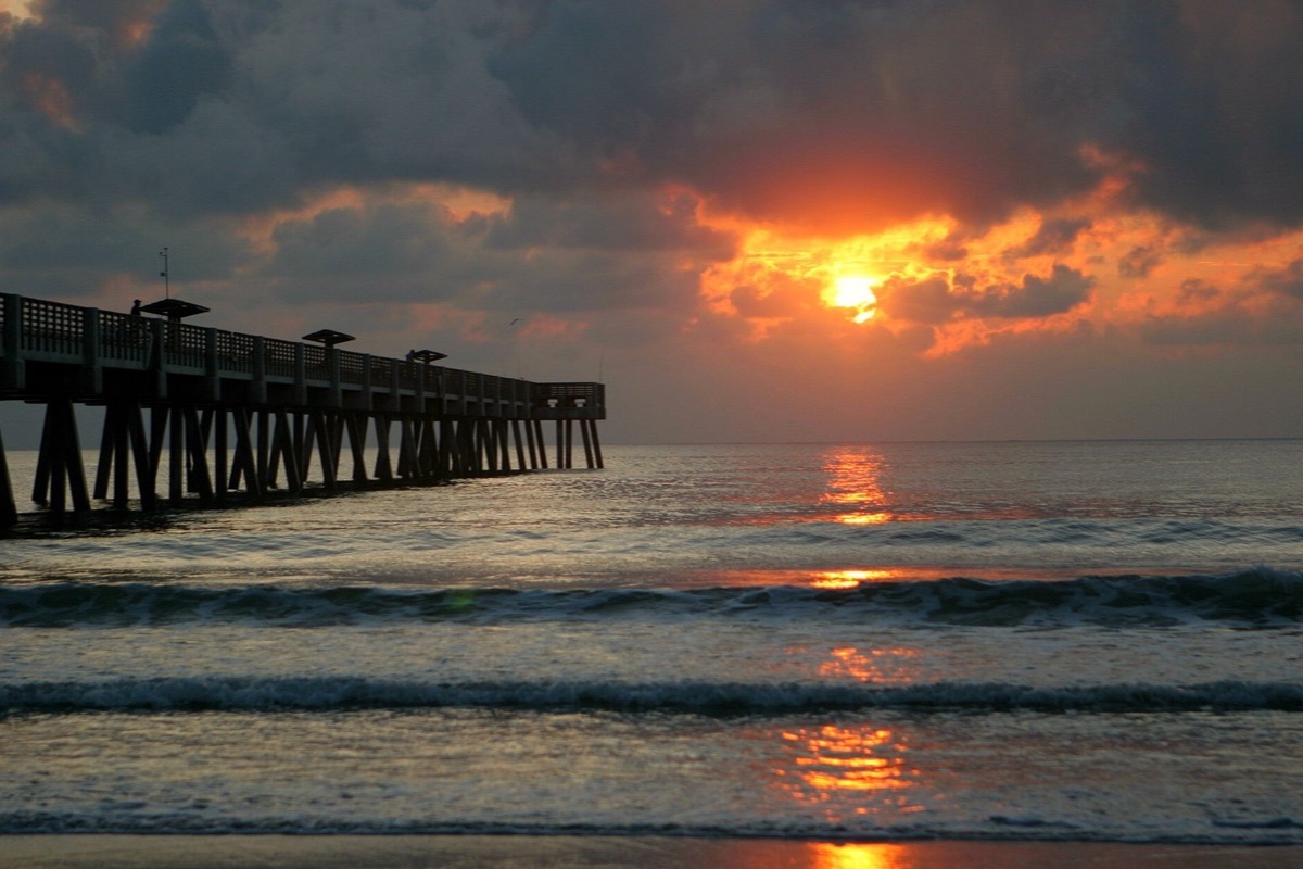 Sunrise over the Atlantic at Jacksonville, the sun breaking through heavy cloud low on the water, its light drawn out across the surf and a long timber pier running out on the left.