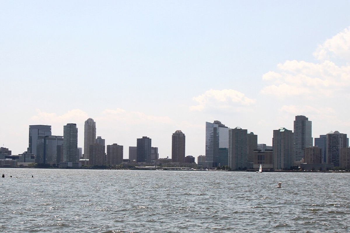 The Jersey City waterfront seen low across the Hudson, a solid line of office and apartment towers along the far bank under a pale sky, with choppy grey water in front.