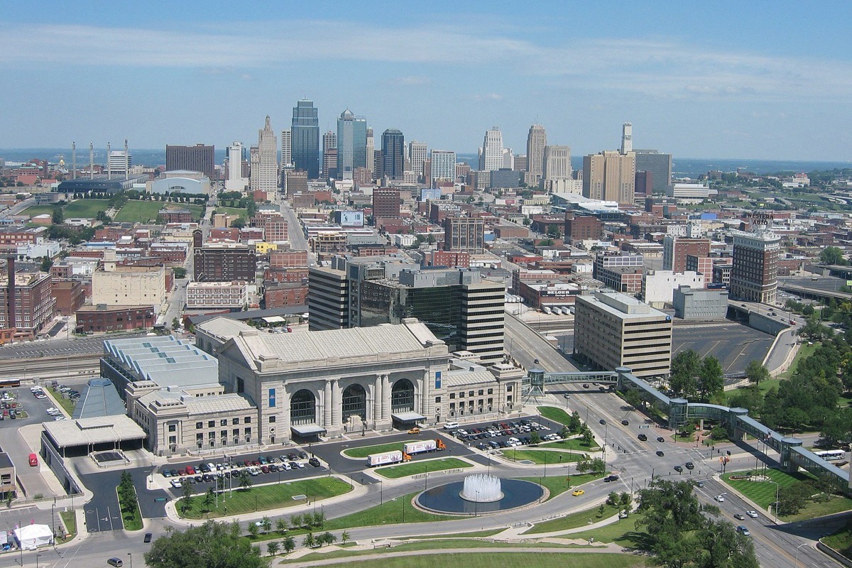 Kansas City from above, the stone mass of the old station and its fountain and lawns in the foreground and the office towers of the city standing behind under a pale summer sky.