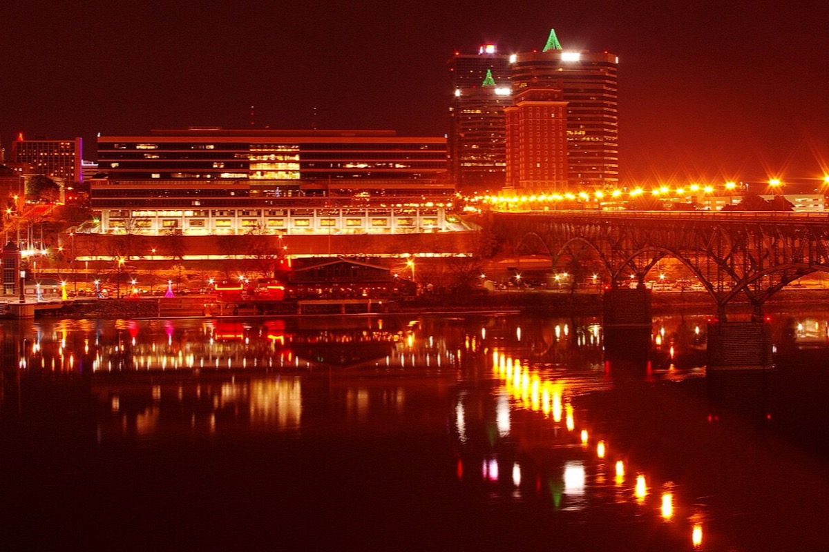 The Knoxville waterfront at night, office towers and a long lit terrace above the river, an arched road bridge on the right, and the lights drawn out in reflections across dark water.