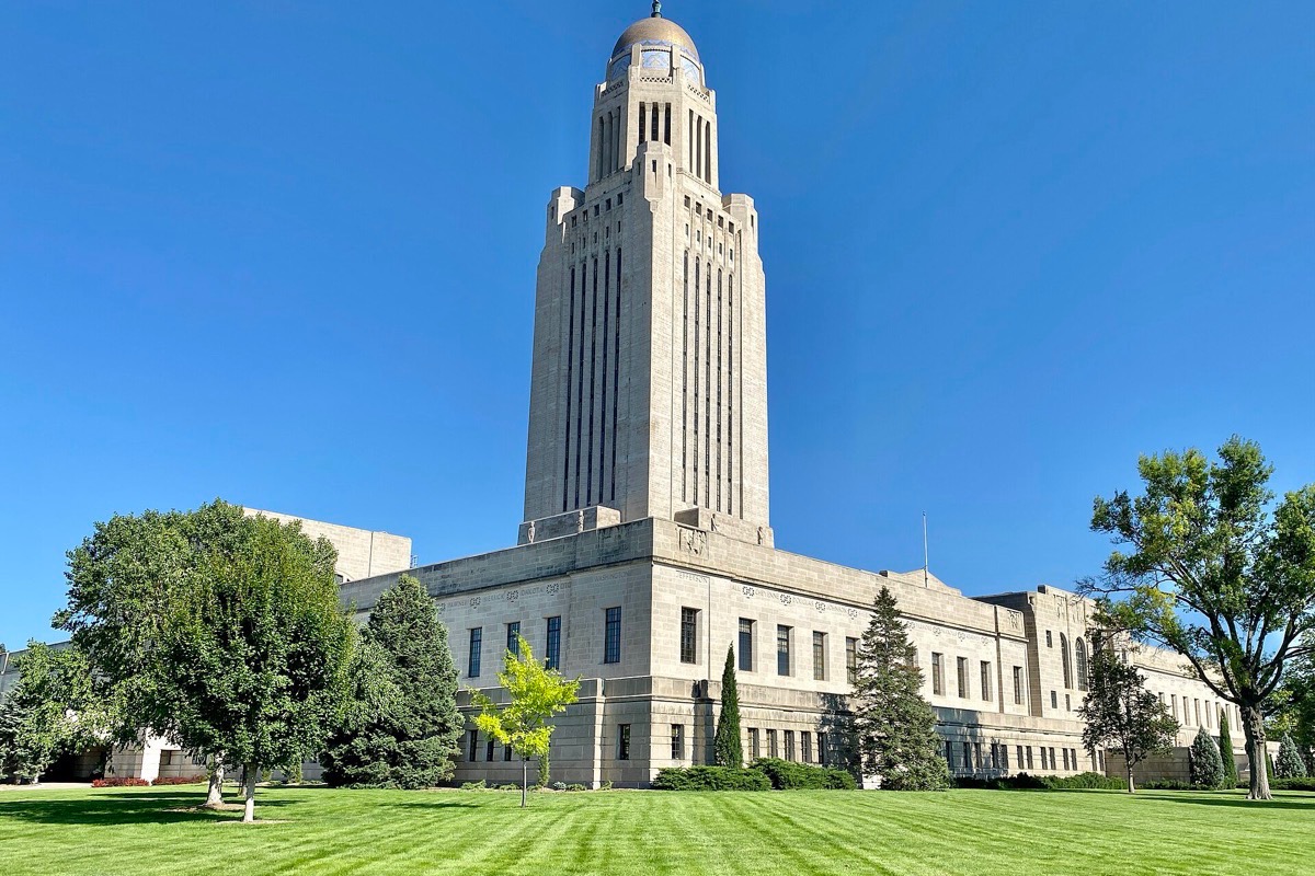 The Nebraska capitol in Lincoln, a tall pale stone tower with a domed top rising from a low square base, standing over mown grass and mature trees under a clear blue sky.