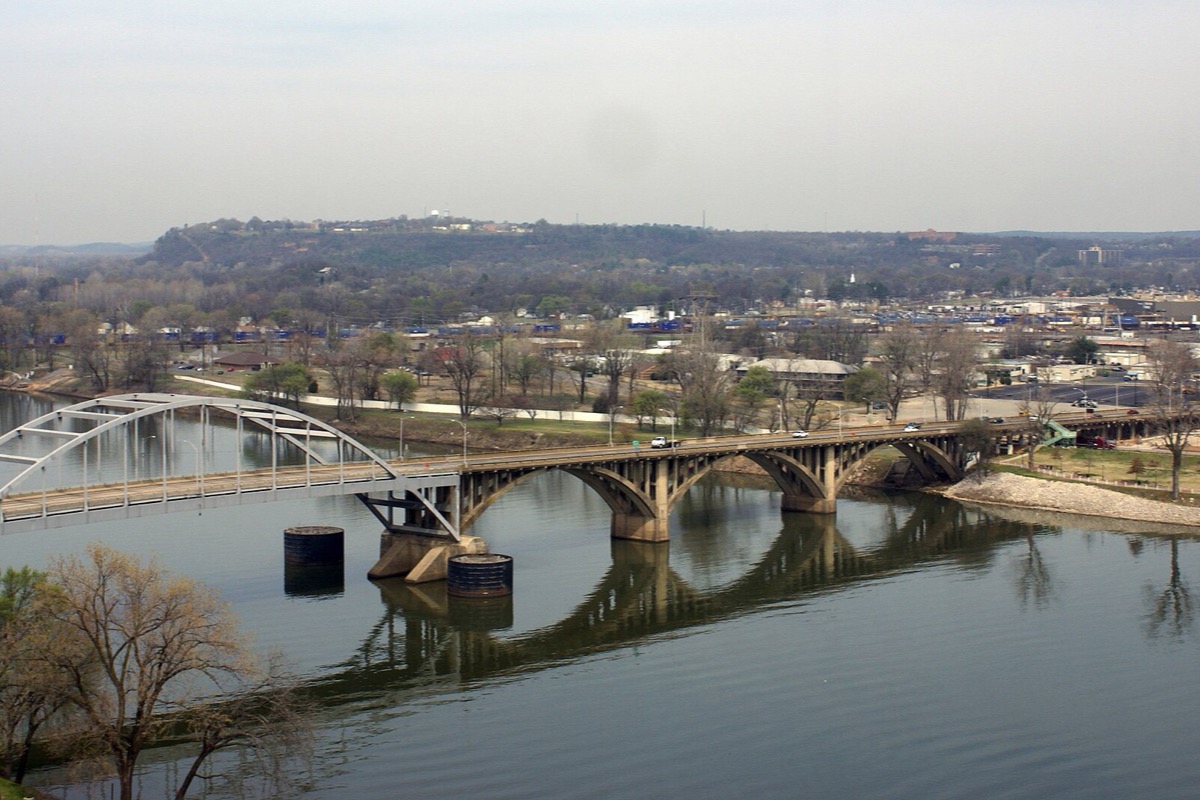 The Arkansas River at Little Rock, with a white steel arch bridge and a row of concrete arch spans crossing calm grey water toward low wooded bluffs and the rooftops of North Little Rock beyond, under a pale overcast sky.