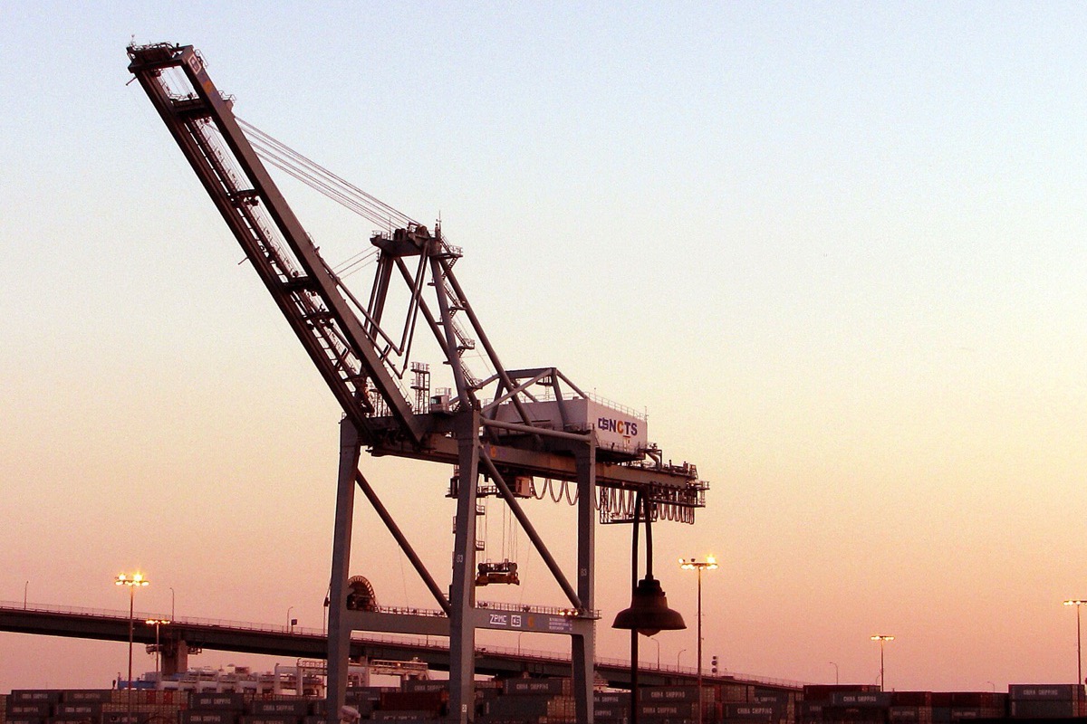 A container crane at the port of Long Beach silhouetted against an orange and blue sunset, its boom raised and stacked containers and harbour lights below it.
